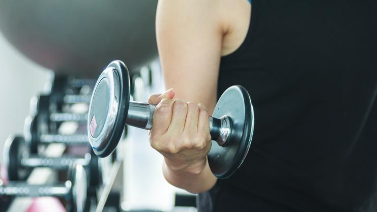 Nahaufnahme einer Hand, die eine Kurzhantel im Fitnessstudio hält, mit weiteren Hanteln im Hintergrund.