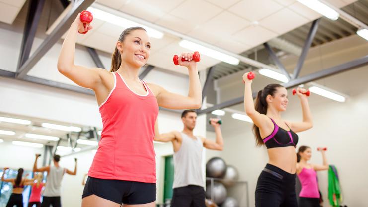 Vier Personen heben Gewichte in einem Fitnessstudio. Zwei Frauen tragen Sporttops, ein Mann hat ein T-Shirt an.