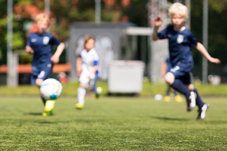 Zwei Kinder in Fußballtrikots rennen auf einem Kunstrasenplatz, ein Ball rollt in der Nähe.