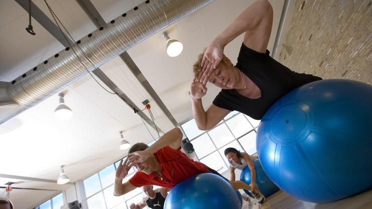 Gruppenfitness im Studio: Teilnehmer üben auf blauen Gymnastikbällen mit Blick zur Fensterfront.