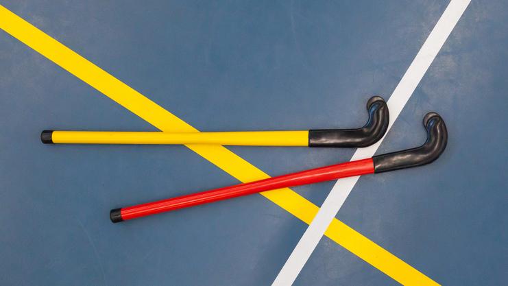 Zwei Hockeyschläger in Gelb und Rot liegen auf einem blauen Sportboden mit weißen und gelben Linien.