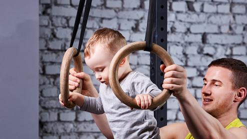 Ein Baby schwingt an Holzturnringen, während ein Erwachsener unterstützend daneben steht. Hintergrund mit Ziegelmauer.