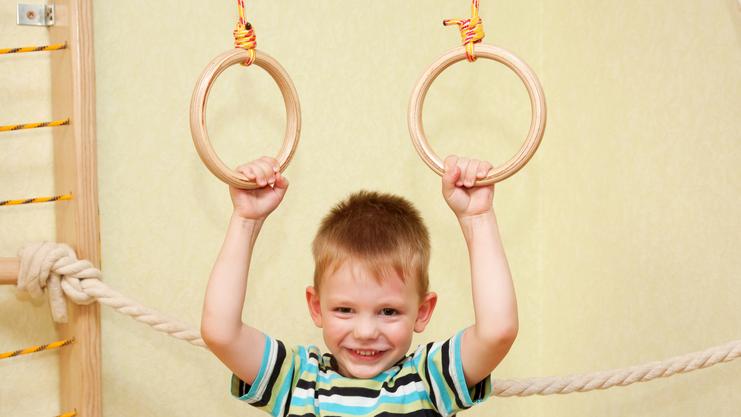 Junge mit einem gestreiften T-Shirt hält sich an zwei Holzringen in einem Spielraum fest und lächelt.
