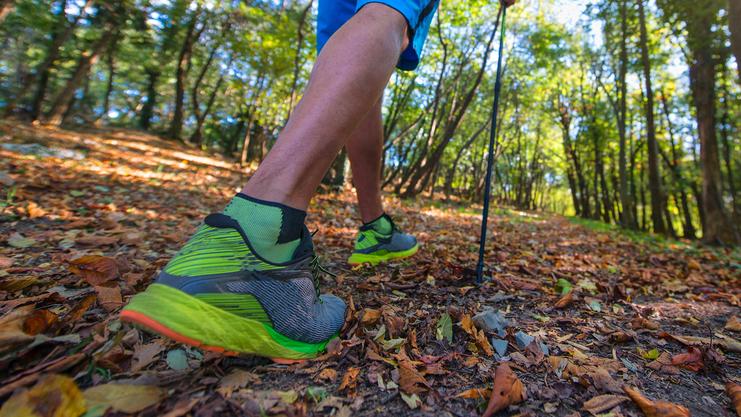 Nahaufnahme eines wandernden Mannes mit Wanderstöcken auf einem mit herbstlichen Blättern bedeckten Waldweg.