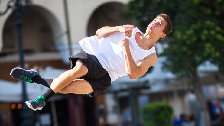 Junger Mann in Sportkleidung springt akrobatisch über einen Bürgersteig in einer sonnigen Stadtlandschaft.