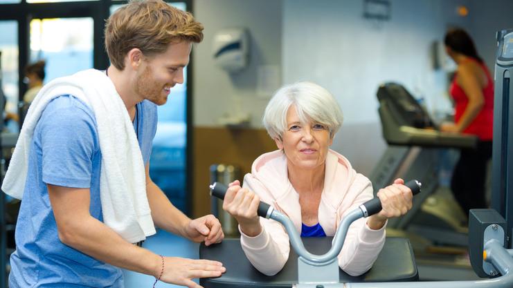 Ältere Frau trainiert mit einem Fitnessgerät, während ein junger Mann sie anleitet und unterstützt.