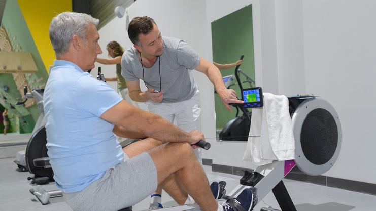Trainer zeigt einem Mann auf einem Rudergerät die Sportdaten auf einem Monitor in einem Fitnessstudio.