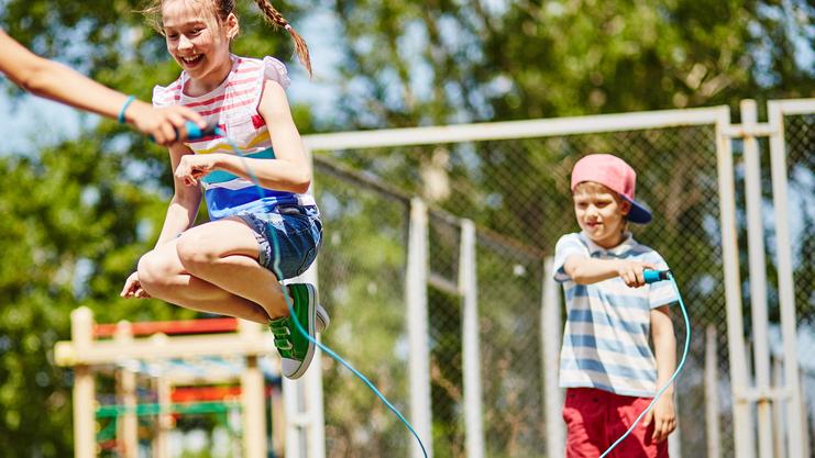 Zwei Kinder spielen im Freien: Ein Mädchen springt über ein Seil, während ein Junge das Seil hält.