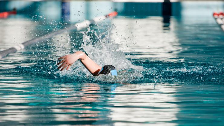 Schwimmerin in Schwarz mit blauer Badekappe zieht eine Bahn im Wasser, spritzt dabei Wasser auf.