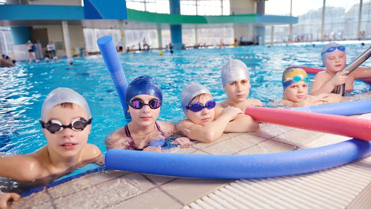 Sechs Kinder im Schwimmbecken lehnen mit Schwimmbrillen und -kappen an der Beckenkante, Schwimmnudeln neben ihnen.