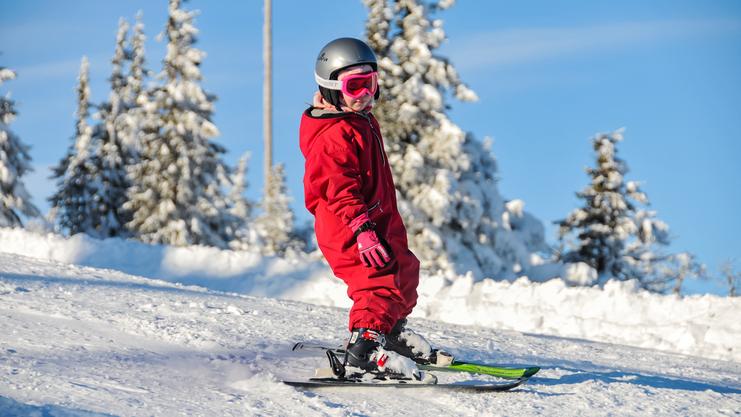 Kind in roter Skikleidung fährt auf einer verschneiten Piste, umgeben von schneebedeckten Bäumen und klarem Himmel.