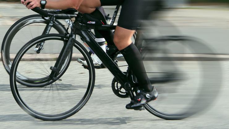 Zwei Radfahrer in sportlichen Outfits fahren schnell auf einer Straße, mit bewegtem Hintergrund und unscharfem Fokus.
