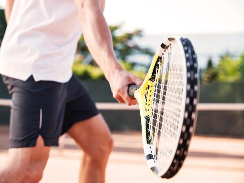 Nahaufnahme eines Tennis-Spielers, der einen Schläger auf einem Sandplatz hält. Hintergrund zeigt Bäume und Himmel.