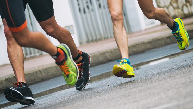 Drei Läufer in Sportkleidung und bunten Schuhen joggen auf einer Stadtstraße entlang einer Wasserpfütze.