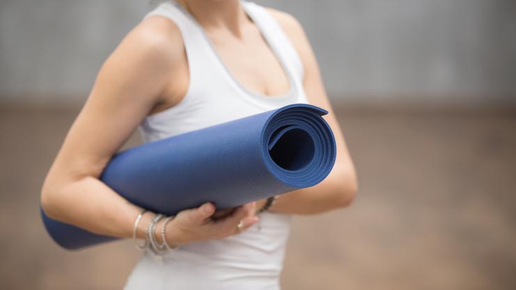 Frau hält eine gerollte blaue Yogamatte und lächelt, sie trägt ein weißes Top in einem Trainingbereich.