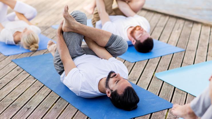 Gruppe von Menschen in Sportkleidung macht Yoga auf blauen Matten auf einem Holzsteig neben einem Gewässer.