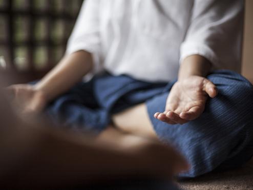 Nahaufnahme einer sitzenden Person in meditativer Haltung mit ausgestreckten Händen auf einem Teppichboden.