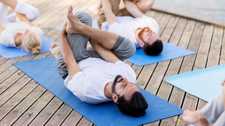 Gruppe von Menschen in Sportkleidung macht Yoga auf blauen Matten auf einem Holzsteig neben einem Gewässer.